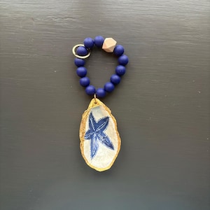 May include: A handcrafted keychain with a blue starfish design on a painted oyster shell pendant. The pendant is suspended from a navy blue beaded bracelet, a gold-tone keyring, and a wooden bead. The background is a dark grey surface.
