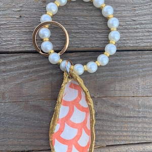 May include: A white pearl keyring with a gold ring and a gold-painted oyster shell charm with a pink and white pattern.