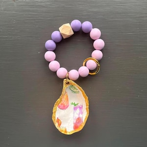 May include: A pink and purple beaded keyring with a gold key ring and a gold oyster shell charm with a painted design of cocktails.