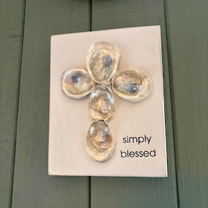 May include: A white wooden sign with a cross made of oyster shells and the text "simply blessed".