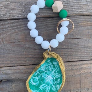 May include: A white beaded keyring with green and wood beads, a gold key ring, and a green and gold oyster shell charm.