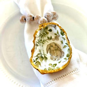 May include: A gold-painted oyster shell napkin ring with a floral and bunny design. The shell is decorated with a white background and green leaves, and a brown bunny is sitting in the center.