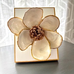 May include: A white flower made from oyster shells with gold accents, centered on a white square canvas with a gold frame. A brown pine cone is in the center of the flower.