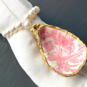 May include: A gold-painted oyster shell napkin ring with a pink floral pattern. The ring is on a white linen napkin.