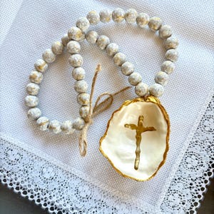 May include: A decorative beaded garland with a white and gold cross-shaped dish. The garland features round, gray beads with gold accents. The dish is white with a gold rim and a gold cross in the center.