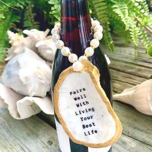May include: A wine bottle with a pearl necklace around the neck and a gold-painted seashell tag that says "Pairs Well With Living Your Best Life".