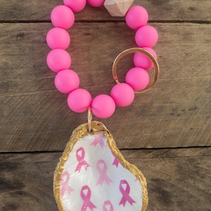 May include: Pink silicone beaded bracelet with a gold key ring and a gold oyster shell charm with pink ribbon pattern.