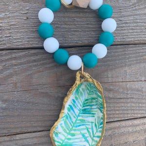 May include: A beaded wristlet keychain with white and teal beads, a gold-coloured ring, and a wooden cube. A painted oyster shell with a gold rim and a green and blue leaf design hangs from the wristlet.