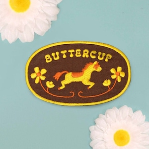 May include: Brown oval embroidered patch with yellow stitching. The patch features a yellow horse with an orange mane and tail, running through a floral design. The text "BUTTERCUP" is embroidered in yellow on the patch.