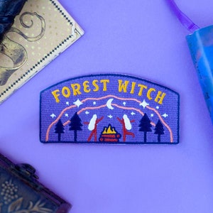 May include: A purple patch with the words "FOREST WITCH" in yellow, featuring a camping scene with trees, a bonfire, and figures. The patch is surrounded by a wand, a bookmark, and a book on a purple surface.