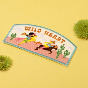 Puede incluir: Un parche bordado blanco con una escena de desierto rosa y naranja. Dos figuras a caballo cabalgan por el desierto. El texto "WILD HEART" está escrito en una fuente retro.