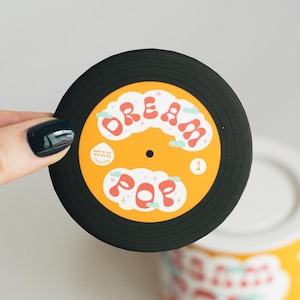 May include: A circular sticker designed to look like a black vinyl record with a yellow centre label. The label features the text "DREAM POP" in red bubble letters, surrounded by small white clouds and stars, with a small number 1 on the right side.