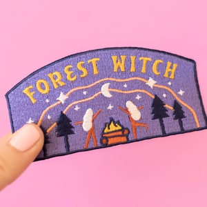 K&ouml;nnte beinhalten: Gestickter Aufn&auml;her mit den Worten "FOREST WITCH" in Gelb auf lila Hintergrund. Der Aufn&auml;her zeigt eine Waldszene mit B&auml;umen, einem Lagerfeuer und tanzenden Figuren unter einem Sternenhimmel.