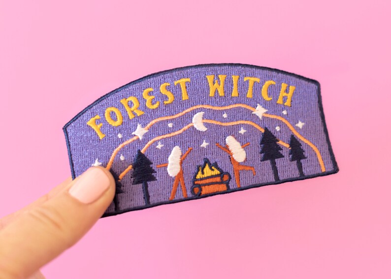 K&ouml;nnte beinhalten: Gestickter Aufn&auml;her mit den Worten "FOREST WITCH" in Gelb auf lila Hintergrund. Der Aufn&auml;her zeigt eine Waldszene mit B&auml;umen, einem Lagerfeuer und tanzenden Figuren unter einem Sternenhimmel.