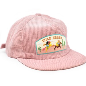 May include: A pink corduroy baseball cap with a patch that says "Wild Heart" and features a cartoon image of two people riding horses in a desert landscape.