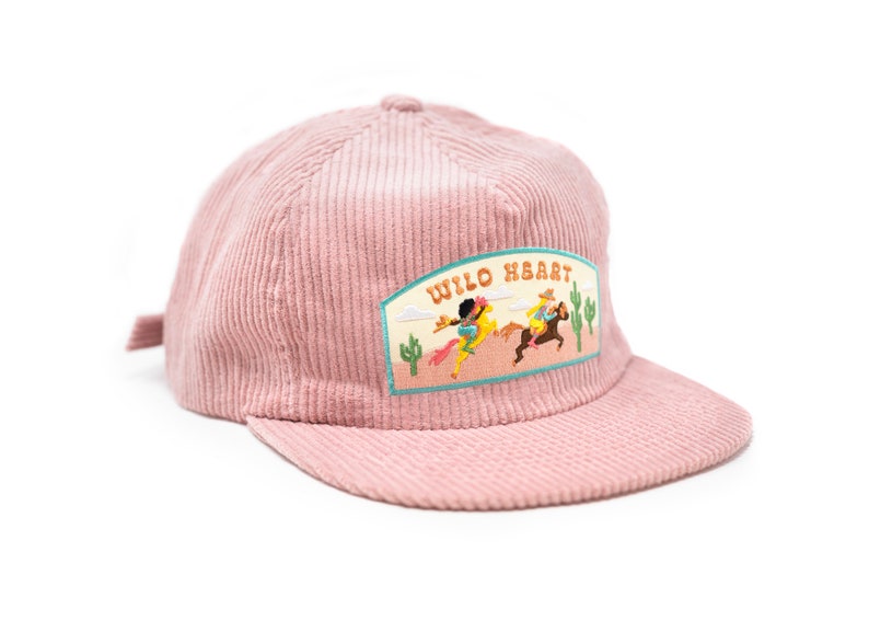 May include: A pink corduroy baseball cap with a patch that says "Wild Heart" and features a cartoon image of two people riding horses in a desert landscape.