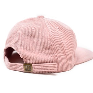 May include: A pink corduroy baseball cap with a brown adjustable strap and a small metal logo on the back.