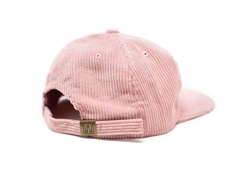May include: A pink corduroy baseball cap with a brown adjustable strap and a small metal logo on the back.