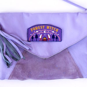 K&ouml;nnte beinhalten: Lavendelfarbene Leder-Clutch mit Klappe und Wildleder-Einsatz. Die Tasche hat eine Quaste und einen Aufn&auml;her mit der Aufschrift "FOREST WITCH" mit einer Campingszene. Sie hat einen Schulterriemen.