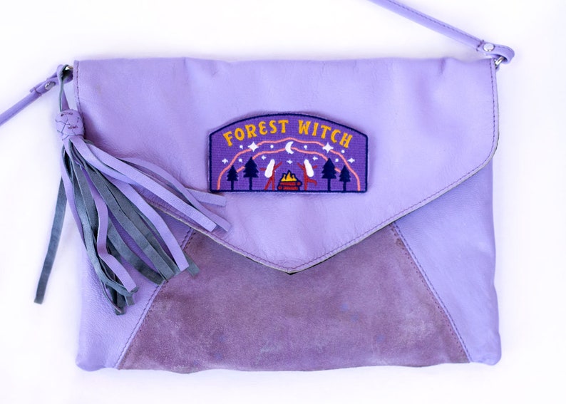 K&ouml;nnte beinhalten: Lavendelfarbene Leder-Clutch mit Klappe und Wildleder-Einsatz. Die Tasche hat eine Quaste und einen Aufn&auml;her mit der Aufschrift "FOREST WITCH" mit einer Campingszene. Sie hat einen Schulterriemen.