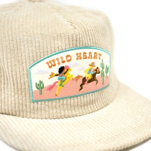 May include: A beige corduroy baseball cap with a patch that says "WILD HEART" and features an embroidered image of two people riding horses in a desert landscape.