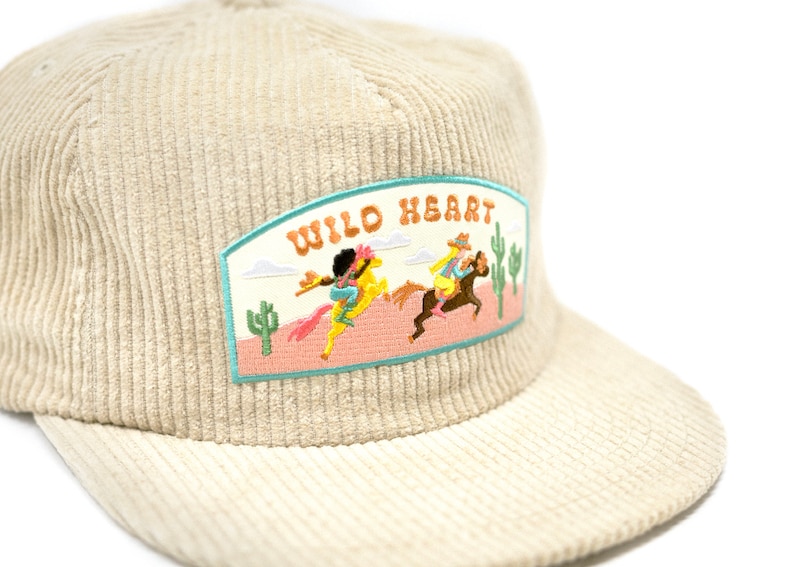 May include: A beige corduroy baseball cap with a patch that says "WILD HEART" and features an embroidered image of two people riding horses in a desert landscape.