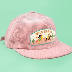 May include: A pink corduroy baseball cap with a patch that says "WILO HEART" and features a cartoon image of two people riding horses in a desert landscape.