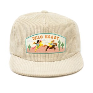 May include: A beige corduroy baseball cap with an embroidered patch that says "WILD HEART" and features a scene of two people riding horses in a desert landscape.