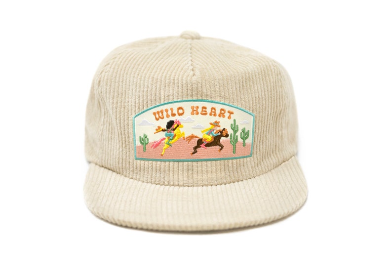 May include: A beige corduroy baseball cap with an embroidered patch that says "WILD HEART" and features a scene of two people riding horses in a desert landscape.
