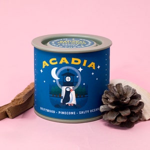 May include: A blue and white candle tin with a lighthouse and a puffin on it. The text on the tin reads "Acadia National Park" and "Driftwood - Pinecone - Salty Ocean".