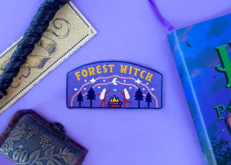 K&ouml;nnte beinhalten: Ein lila Aufn&auml;her mit den gelben Worten "FOREST WITCH", der eine Campingszene mit B&auml;umen, einem Lagerfeuer und Figuren zeigt. Der Aufn&auml;her befindet sich neben einem Zauberstab, einem Lesezeichen und einem Buch auf einer lila Oberfl&auml;che.