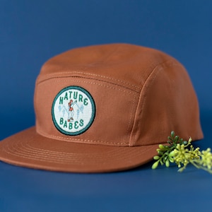 May include: A brown 5-panel cap with a green and white patch that says "NATURE BABES" and features a cartoon woman holding an ax.