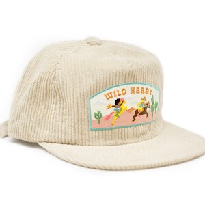 May include: A beige corduroy baseball cap with an embroidered patch that says "Wild Heart" and features a scene of two people riding horses in a desert landscape.