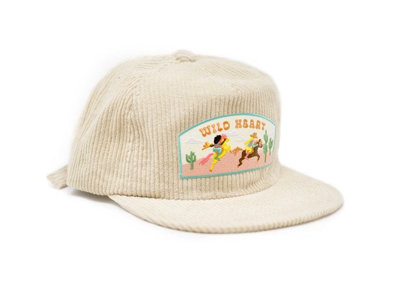 May include: A beige corduroy baseball cap with an embroidered patch that says "Wild Heart" and features a scene of two people riding horses in a desert landscape.