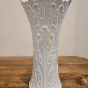 May include: A white ceramic vase with a flared top and a detailed leaf pattern. The vase has a round base and stands approximately 10 inches tall. The design features raised, curved leaf shapes that extend from the base to the top.