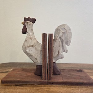 May include: Wooden rooster bookends. The bookends feature a carved rooster design with a white body, brown comb, and brown feet. The bookends are mounted on a rectangular wooden base. The rooster is facing left.