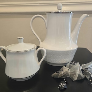May include: White porcelain tea set with silver trim, including a teapot, sugar bowl, and tea bags. The teapot has a textured lower half. The tea bags have the text "TRUE TEA".