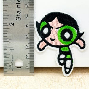 Op de afbeelding: Geborduurde strijk-op patch van een cartoon meisje met groene ogen en een groen-zwarte outfit, in de kleuren groen, zwart en roze.