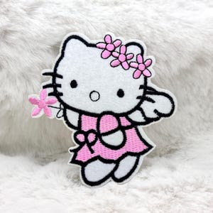 Puede incluir: Parche bordado blanco con un gato de dibujos animados con vestido rosa y corona de flores. El gato sostiene una flor rosa. El parche tiene un contorno negro sobre un fondo blanco y esponjoso.