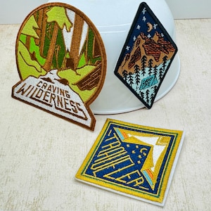 Pode incluir: Três patches bordados com temas ao ar livre. Um é oval com uma cena de floresta e o texto "Craving Wilderness". Outro é em forma de diamante com uma cena de montanha e o texto "Camp Life". O terceiro é quadrado com o texto "Thrill Seeker".