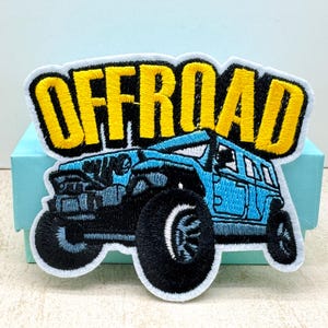 May include: Embroidered patch showing a blue off-road vehicle with large black tyres. The word "OFFROAD" is embroidered in yellow block letters above the vehicle. The patch has a white border, suitable for clothing or accessories.