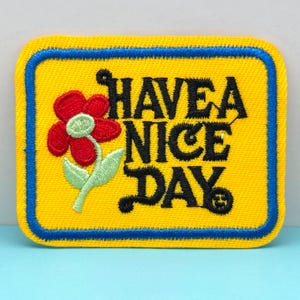 May include: A yellow patch with a blue border and the words "HAVE A NICE DAY" in black. A red flower with green leaves is on the left side of the patch. The patch is rectangular with rounded corners.
