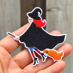 Puede incluir: Un parche bordado que muestra a una bruja con cabello rojo brillante, un sombrero puntiagudo negro y una capa negra ondeante. La bruja monta una escoba con cerdas naranjas y lleva leggings azules con cordones rojos y botas negras.