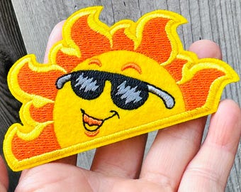 Op de afbeelding: Geborduurde patch met een lachende gele zon met een zwarte zonnebril. De zon heeft oranje vlammen rondom zijn gezicht. De patch is waarschijnlijk voor kleding of accessoires.