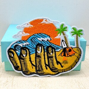 Puede incluir: Parche bordado que representa una escena de playa con una puesta de sol, olas del océano, palmeras y una tienda de campaña. La palabra "WAVE" está bordada en amarillo y negro. El parche está sobre una superficie azul claro.