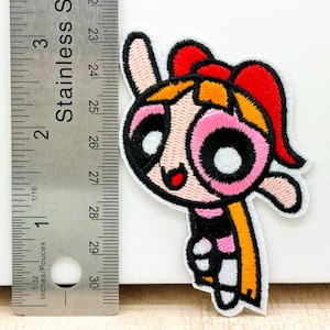 Op de afbeelding: Een geborduurde strijk-patch met een cartoonfiguur met roze wangen, zwarte ogen en een geel en rood hoofdband. Het personage draagt een zwart-roze gestreept shirt en een gele sjaal.