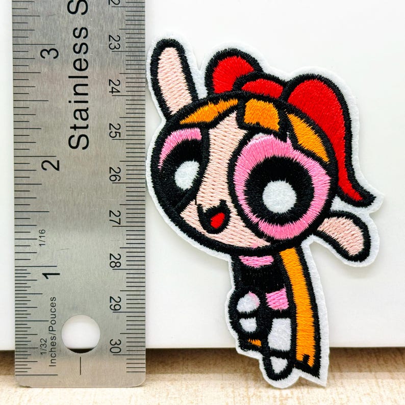 Op de afbeelding: Een geborduurde strijk-patch met een cartoonfiguur met roze wangen, zwarte ogen en een geel en rood hoofdband. Het personage draagt een zwart-roze gestreept shirt en een gele sjaal.