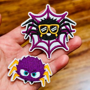 May include: Two embroidered spider patches. One black spider with yellow sunglasses and a purple web. The other is a purple spider with blue eyes and yellow lightning bolt accents. Both have white borders.