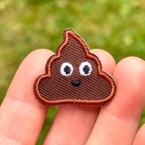 May include: A brown embroidered patch in the shape of a smiling poop emoji. The patch has a brown outline and features white eyes and a small black mouth. The background is a blurred green.