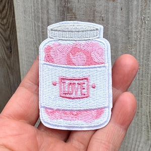 May include: Embroidered patch featuring a jar filled with pink hearts. The jar has a white label with the word "LOVE" in pink. The patch is light blue with a gray lid.